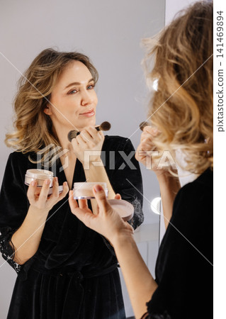 Beauty routine. Face makeup. Woman applying cosmetic product powder with brush for treatment using mirror at modern home. Beauty routine. Face makeup. Woman applying cosmetic product powder with brush for treatment using mirror at modern home. 141469894