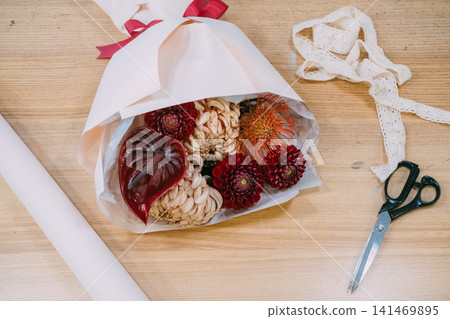 Flower craft. Bouquet preparing. Red dahlias chrysanthemums color anthurium protea wrapped in paper ribbon on wooden table in professional florist shop. 141469895