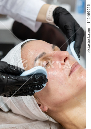 Woman product removing. Skin treatment. Professional cosmetologist hands in gloves with facial mask for client using cosmetic sponge in spa salon. Woman product removing. Skin treatment. Professional cosmetologist hands in gloves with facial mask for client using cosmetic sponge in spa salon. 141469926