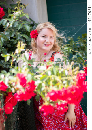 Adult blonde woman smiles with a red rose tucked into her hair, framed by softly blurred blossoms and leafy branches outdoors. Adult blonde woman smiles with a red rose tucked into her hair, framed by softly blurred blossoms and leafy branches outdoors. 141470814