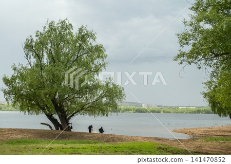 Fishermen on sandy riverbank under tree on cloudy day Fishermen on sandy riverbank under tree on cloudy day 141470852