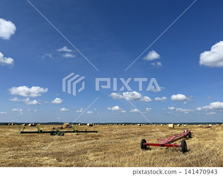 Farm trailers in harvested field with round hay bales under blue summer sky and rural landscape Farm trailers in harvested field with round hay bales under blue summer sky and rural landscape 141470943
