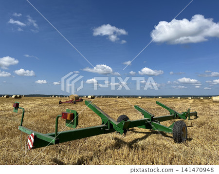 Green header trailer awaiting transport in harvested field with round hay bales under summer sky 141470948