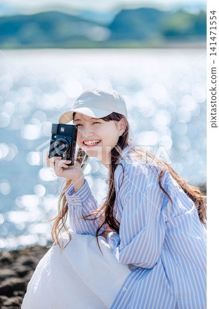 A woman in a light blue shirt holds a film camera by the sparkling lakeside. 141471554