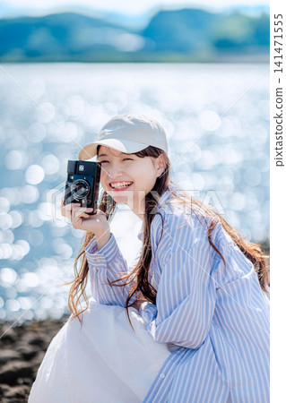 A woman in a white hat smiles, holding a camera by the water's edge. A woman in a white hat smiles, holding a camera by the water's edge. 141471555