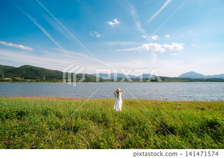 A woman in a white dress stands on a lakeside meadow under the summer sky. A woman in a white dress stands on a lakeside meadow under the summer sky. 141471574