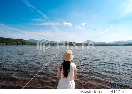 The back view of a woman in a white dress, wearing a straw hat, gazing at the lake. The back view of a woman in a white dress, wearing a straw hat, gazing at the lake. 141471603
