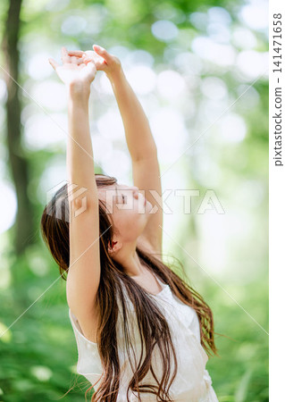 A woman stretches her arms and refreshes herself in a forest dappled with sunlight. A woman stretches her arms and refreshes herself in a forest dappled with sunlight. 141471658