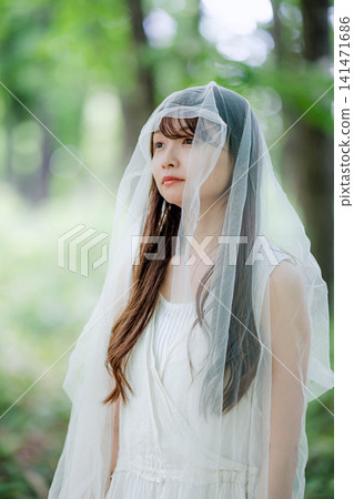 陽光斑駁的森林裡,一位身穿白色洋裝、頭戴面紗的女子。 陽光斑駁的森林裡,一位身穿白色洋裝、頭戴面紗的女子。 141471686