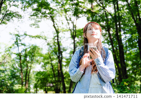A woman in jeans looks up at her smartphone in a lush green forest. A woman in jeans looks up at her smartphone in a lush green forest. 141471711