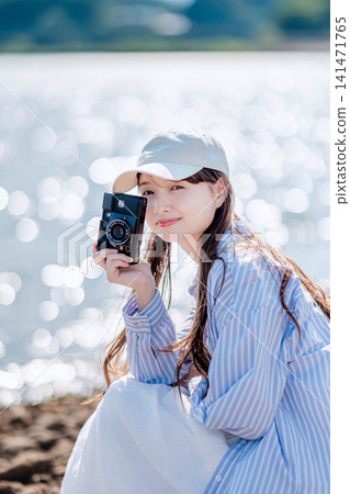 A woman in a white hat holds a camera by the sparkling lakeside. A woman in a white hat holds a camera by the sparkling lakeside. 141471765