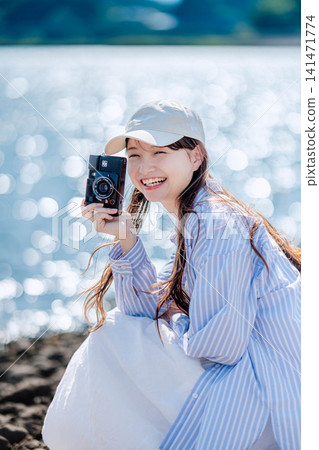 一位身穿淺藍色襯衫的女子坐在湖邊,手裡拿著相機。 一位身穿淺藍色襯衫的女子坐在湖邊,手裡拿著相機。 141471774