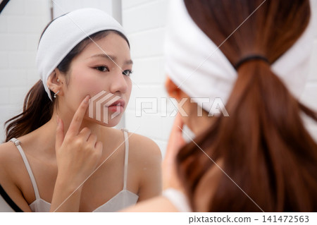 Beautiful young asian woman examining skin of facial while looking mirror in bathroom at home. Beautiful young asian woman examining skin of facial while looking mirror in bathroom at home. 141472563