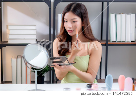 Happy young asian woman applying makeup with a brush on cheek looking mirror. Happy young asian woman applying makeup with a brush on cheek looking mirror. 141472595