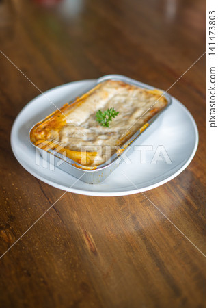 Fresh baked lasagna or macaroni and cheese in aluminum baking tray on white plate, creamy melted cheese comfort food served on rustic wooden table with copy space Fresh baked lasagna or macaroni and cheese in aluminum baking tray on white plate, creamy melted cheese comfort food served on rustic wooden table with copy space 141473803