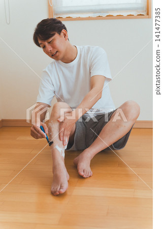 A man in his 40s using a T-shaped razor to remove his leg hair. A man in his 40s using a T-shaped razor to remove his leg hair. 141477385