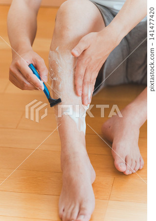 A man shaving his leg hair with a T-shaped razor. A man shaving his leg hair with a T-shaped razor. 141477420