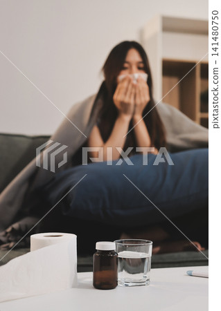 sick asian young woman sneezing into tissue paper while covered with a blanket and sitting on sofa in the living room sick asian young woman sneezing into tissue paper while covered with a blanket and sitting on sofa in the living room 141480750