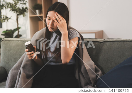 sick asian young woman sneezing into tissue paper while covered with a blanket and sitting on sofa in the living room sick asian young woman sneezing into tissue paper while covered with a blanket and sitting on sofa in the living room 141480755