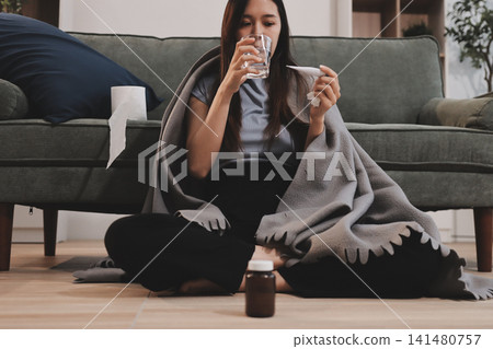 sick asian young woman sneezing into tissue paper while covered with a blanket and sitting on sofa in the living room sick asian young woman sneezing into tissue paper while covered with a blanket and sitting on sofa in the living room 141480757