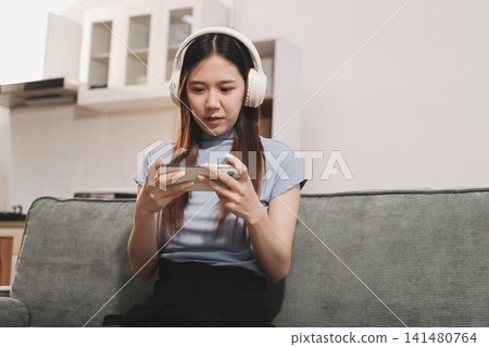 Beautiful young asian woman sitting on sofa and holding joystick playing online video game in living room at house Beautiful young asian woman sitting on sofa and holding joystick playing online video game in living room at house 141480764