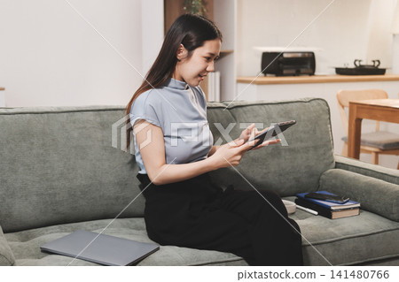 Freelance Asian woman holding mobile phone typing on laptop and working from home. Happy young girl sitting on sofa in living room. Freelance Asian woman holding mobile phone typing on laptop and working from home. Happy young girl sitting on sofa in living room. 141480766