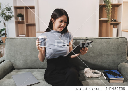 Freelance Asian woman holding mobile phone typing on laptop and working from home. Happy young girl sitting on sofa in living room. 141480768