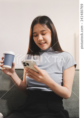 Freelance Asian woman holding mobile phone typing on laptop and working from home. Happy young girl sitting on sofa in living room. 141480769