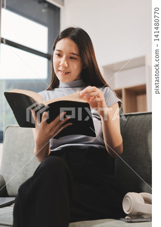Freelance Asian woman holding mobile phone typing on laptop and working from home. Happy young girl sitting on sofa in living room. 141480770