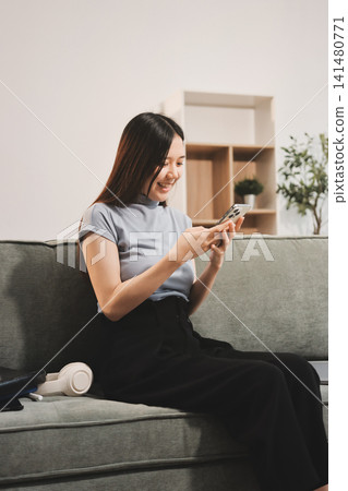 Freelance Asian woman holding mobile phone typing on laptop and working from home. Happy young girl sitting on sofa in living room. 141480771