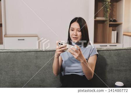 Freelance Asian woman holding mobile phone typing on laptop and working from home. Happy young girl sitting on sofa in living room. 141480772