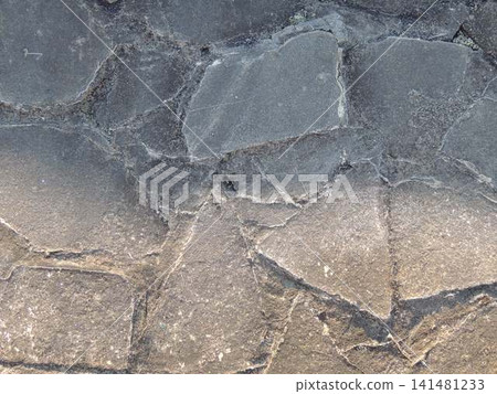 A close-up of an old pavement made of flat, irregularly shaped gray stones is shown. The large elements are bonded with cement mortar, and cracks, chips, and a rough texture are visible on the surface A close-up of an old pavement made of flat, irregularly shaped gray stones is shown. The large elements are bonded with cement mortar, and cracks, chips, and a rough texture are visible on the surface 141481233
