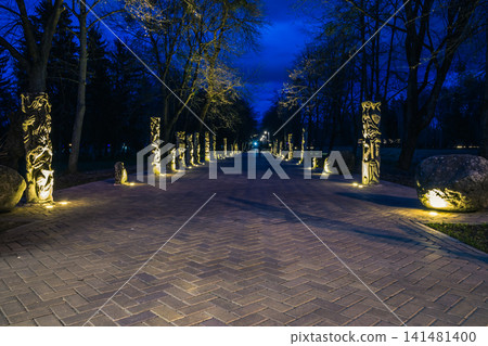 The tiled road in the night park with stone idols illuminated by lamps. Stone statues in the park at night. Illumination of a park road with lanterns at night. Lutsk The tiled road in the night park with stone idols illuminated by lamps. Stone statues in the park at night. Illumination of a park road with lanterns at night. Lutsk 141481400