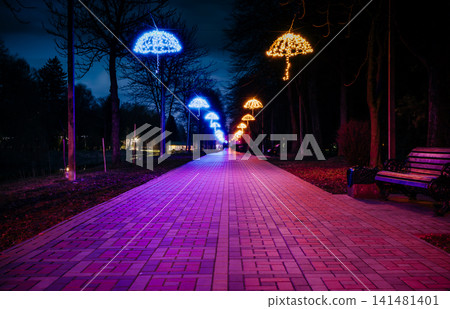 The tiled road in the night park with illuminated umbrellas. Benches in the park at night. Illumination of a park road with small lanterns on umbrellas suspended above the path at night. Lutsk.Ukraine The tiled road in the night park with illuminated umbrellas. Benches in the park at night. Illumination of a park road with small lanterns on umbrellas suspended above the path at night. Lutsk.Ukraine 141481401