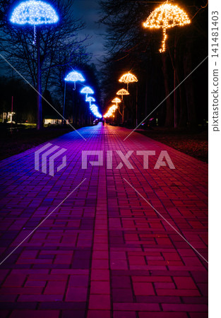 The tiled road in the night park with illuminated umbrellas. Benches in the park at night. Illumination of a park road with small lanterns on umbrellas suspended above the path at night. Lutsk.Ukraine The tiled road in the night park with illuminated umbrellas. Benches in the park at night. Illumination of a park road with small lanterns on umbrellas suspended above the path at night. Lutsk.Ukraine 141481403