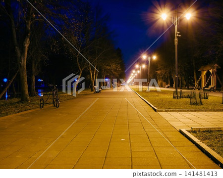 Night city park. Wooden benches, street lights and park alley. The tiled road in the night park with lanterns. Benches in the park at night. Illumination of a park road with lanterns at night. Lutsk Night city park. Wooden benches, street lights and park alley. The tiled road in the night park with lanterns. Benches in the park at night. Illumination of a park road with lanterns at night. Lutsk 141481413
