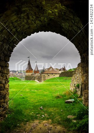View of the Old Kamyanets-Podilsky castle from under an old arch under a cloudy gray sky. The fortress is located amidst picturesque nature in the historical city of Kamyanets-Podilsky, Ukraine. View of the Old Kamyanets-Podilsky castle from under an old arch under a cloudy gray sky. The fortress is located amidst picturesque nature in the historical city of Kamyanets-Podilsky, Ukraine. 141482234