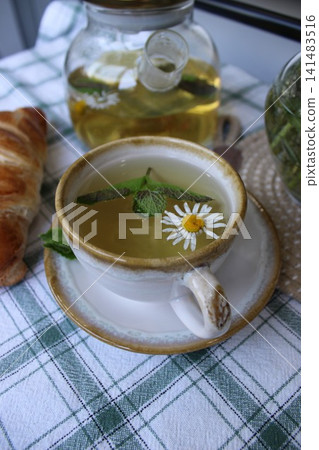 Herbal tea in a decorative cup with mint leaves and a daisy flower, accompanied by a croissant on a checkered tablecloth in a cozy kitchen setting 141483516