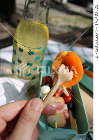 Hand holding a small fork with an apricot slice above a colorful picnic box filled with assorted fruits and a bottle of lemonade on a checkered tablecloth Hand holding a small fork with an apricot slice above a colorful picnic box filled with assorted fruits and a bottle of lemonade on a checkered tablecloth 141483529