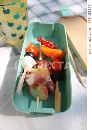 Colorful skewers with meat, cheese, and fruits arranged in a turquoise serving tray beside a glass of beverage on a picnic blanket outdoors Colorful skewers with meat, cheese, and fruits arranged in a turquoise serving tray beside a glass of beverage on a picnic blanket outdoors 141483531