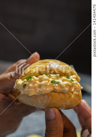 Japanese tamago sando with creamy egg filling and herbs held by hand in warm rustic cafe atmosphere. Asian comfort food concept. Japanese tamago sando with creamy egg filling and herbs held by hand in warm rustic cafe atmosphere. Asian comfort food concept. 141484479