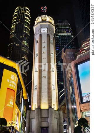 The illuminated People's Liberation Monument glows brightly at night in the center of Chongqing's vibrant commercial district. Towering modern skyscrapers and brightly lit storefronts surround the The illuminated People's Liberation Monument glows brightly at night in the center of Chongqing's vibrant commercial district. Towering modern skyscrapers and brightly lit storefronts surround the 141484537
