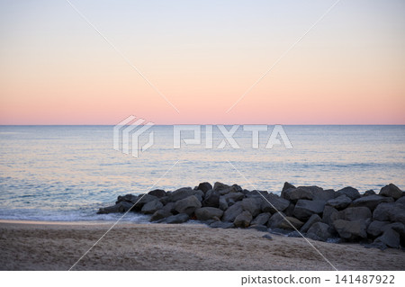 Rocks Along A Sandy Shoreline Under A Pastel Sunset Sky Rocks Along A Sandy Shoreline Under A Pastel Sunset Sky 141487922