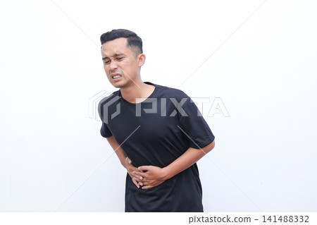 Man grips abdomen, grimacing in discomfort. His body language conveys sudden pain. White background isolates the subject clearly. Ideal for health, medical, or wellness topics Man grips abdomen, grimacing in discomfort. His body language conveys sudden pain. White background isolates the subject clearly. Ideal for health, medical, or wellness topics 141488332