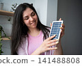 Smiling woman holding a small solar panel indoors near a window. Green technology sustainability and alternative energy choice 141488808