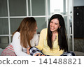 Two women laughing together on a bed in a cozy bedroom setting. Friendship sisterhood family bonding and emotional support 141488821