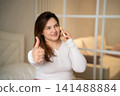 Woman giving a thumbs up while talking on a smartphone in a cozy room. Positive communication online feedback and remote consultation approval 141488884