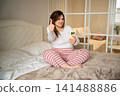 Woman sitting on a bed in a cozy bedroom giving a thumbs up with a smartphone. Home lifestyle weekend self-care and bedroom leisure 141488886