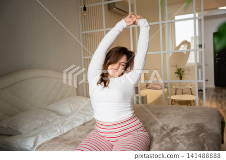 Woman stretching on bed in cozy bedroom during morning routine. Wellness and healthy wake-up habits for improved sleep quality Woman stretching on bed in cozy bedroom during morning routine. Wellness and healthy wake-up habits for improved sleep quality 141488888