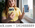 Woman using smartphone connected to portable solar charger by a window. Clean energy mobile connectivity and renewable technology 141488889